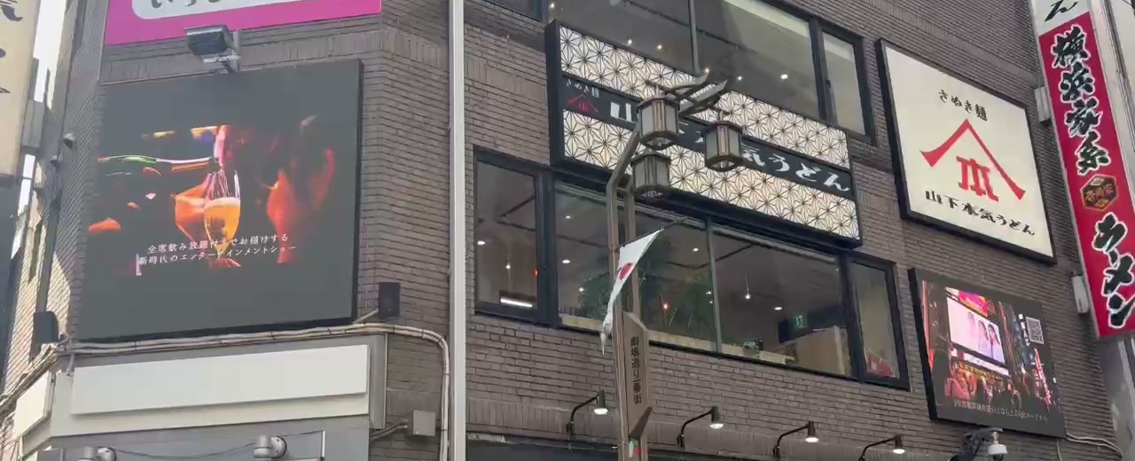繁華街のビル壁面に取り付けられた横長のビジョンに、暗い背景の映像が流れている様子。周囲には飲食店の看板が並んでいる