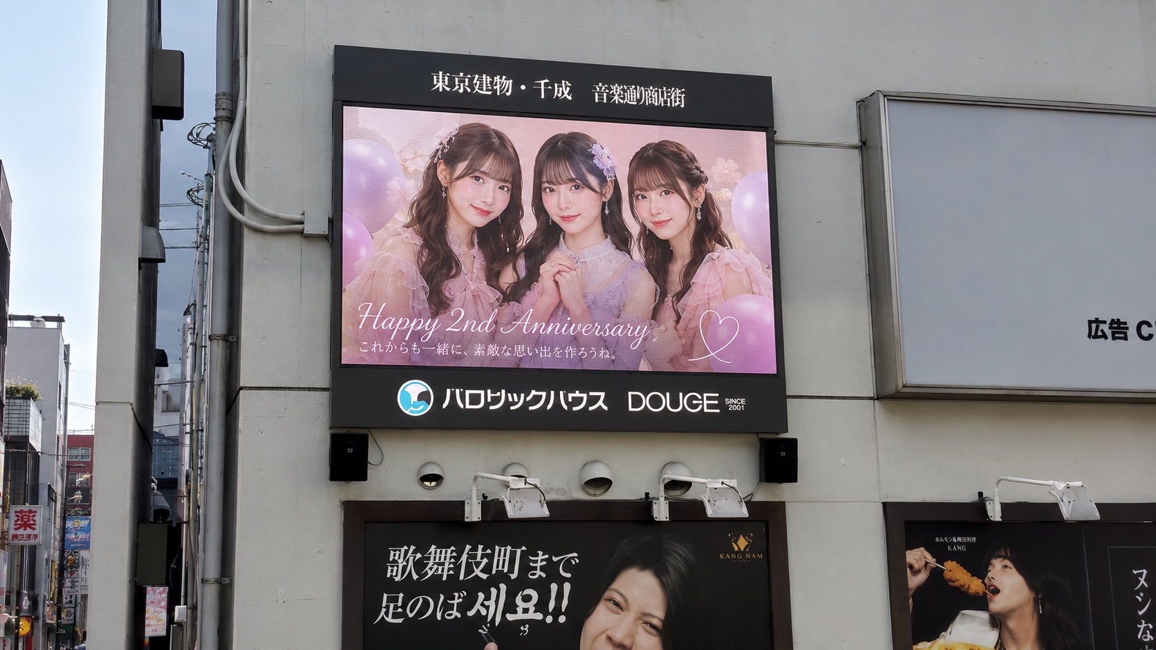 商店街のビル壁面ビジョンに、女性3人組の写真と「Happy 2nd Anniversary」「これからも一緒に、素敵な思い出を作ろうね。」の文字が入った応援広告が映っている放映イメージ